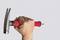 A hand is holding a hammer with a red handle and is hammering a nail into the wall. The background is grey.