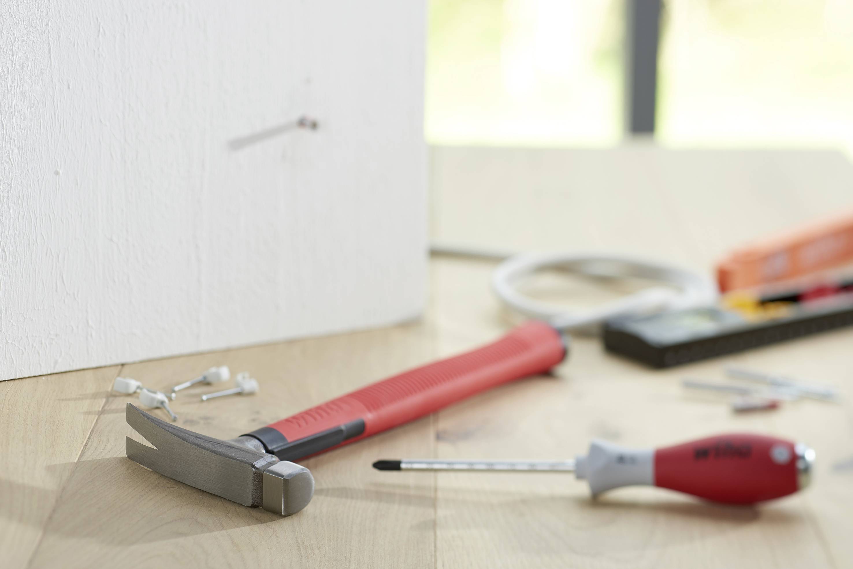 A hammer is lying on a wooden table in front of a wall with a protruding nail. Screwdrivers and screws are visible in the background.