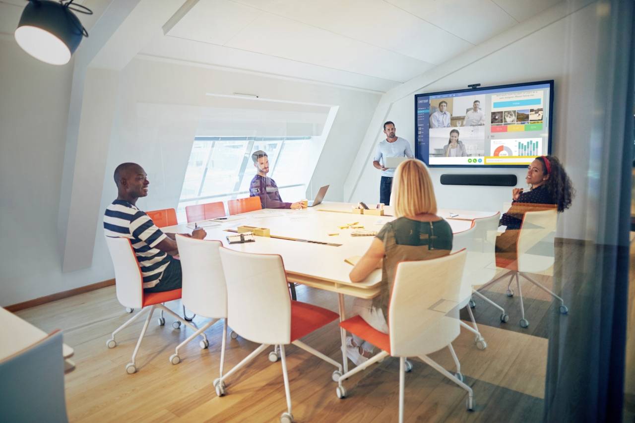 A group of people in a modern conference room are holding a meeting. A man is presenting on a screen while others sit and listen.
