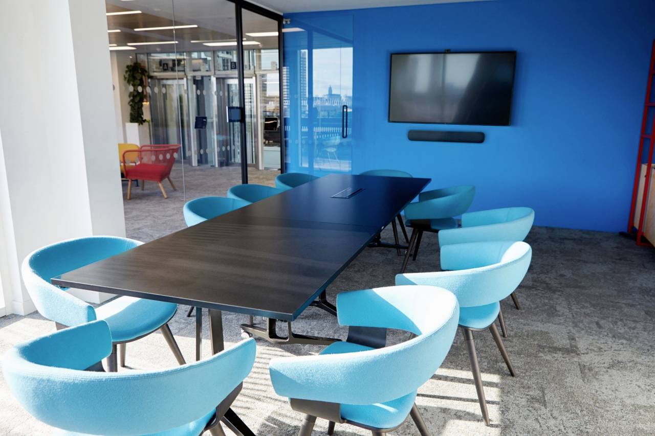 A modern meeting room with a long wooden table, surrounded by blue chairs. A screen is hanging on the blue wall.