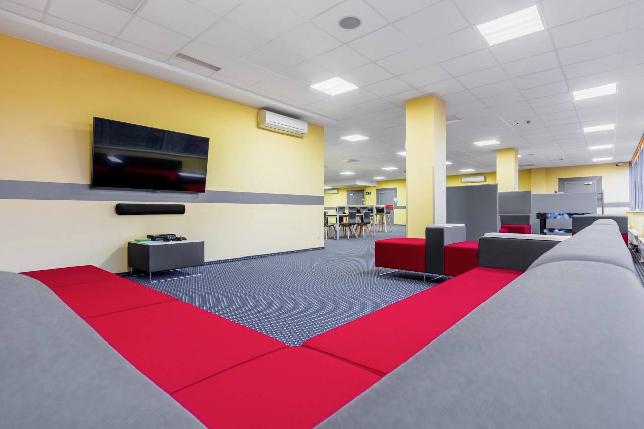 A modern conference room with yellow walls, grey and red sofas, a large television mounted on the wall, and a table in the background.