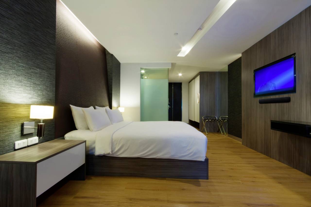 A modern hotel room with a large bed, white pillows, bedside lamps, wooden flooring, and a flat-screen television mounted on the wall.