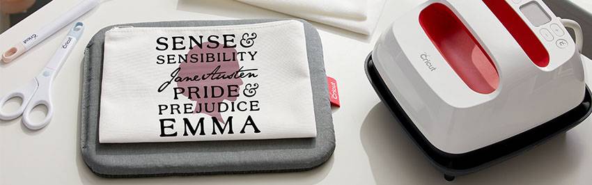 A bag containing 'Sense & Sensibility', 'Pride & Prejudice', 'Emma' lies on a heating mat next to a heat press device and scissors.