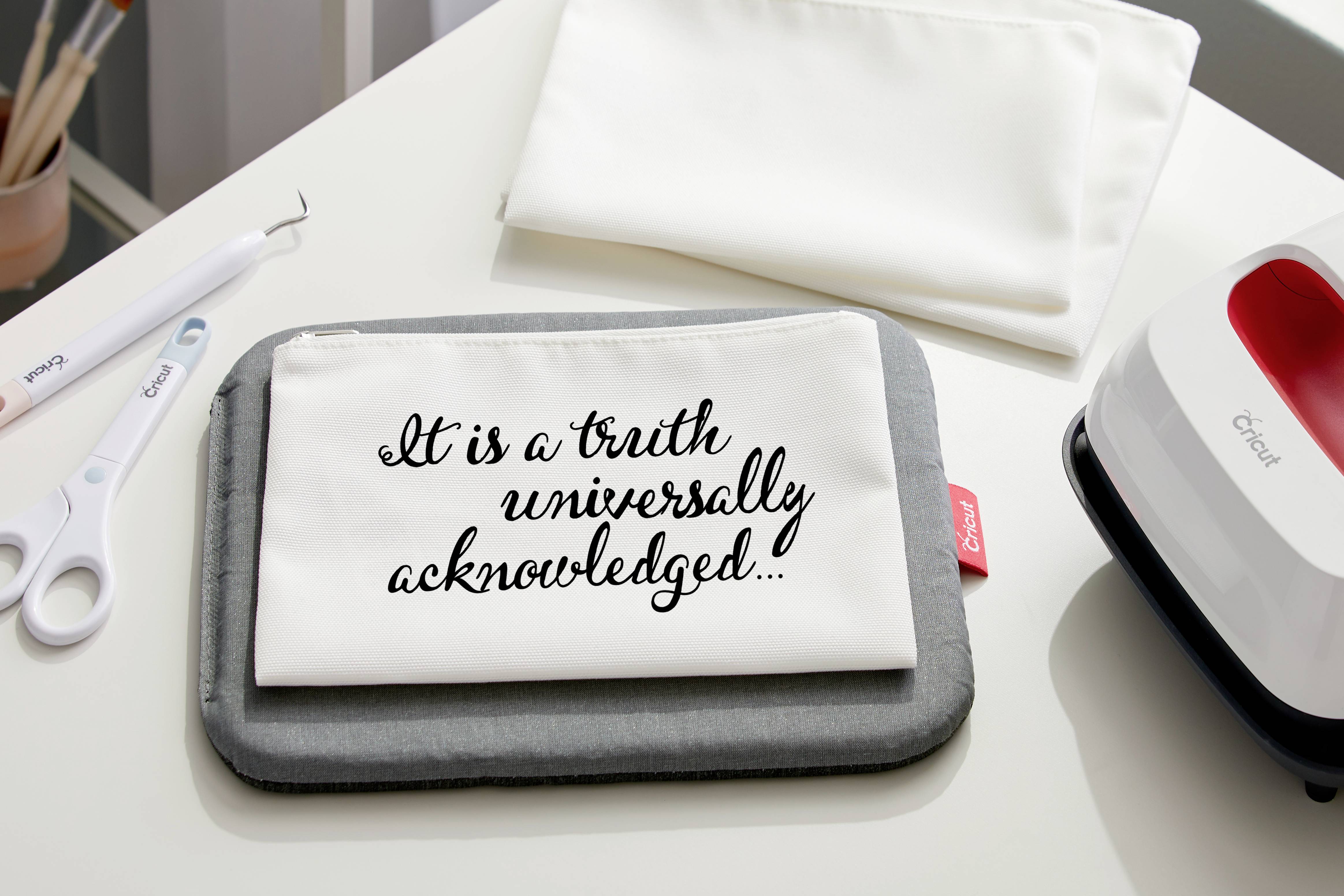 White fabric tote bag with the text 'It is a truth universally acknowledged...' on a table with craft tools and a heat press.