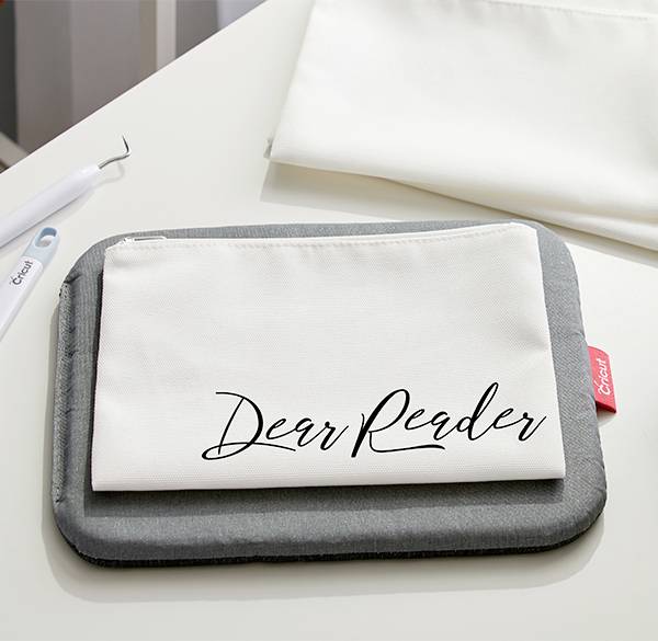 Beige bag with black 'Dear Reader' lettering on a table, next to a plain grey case and a tool.