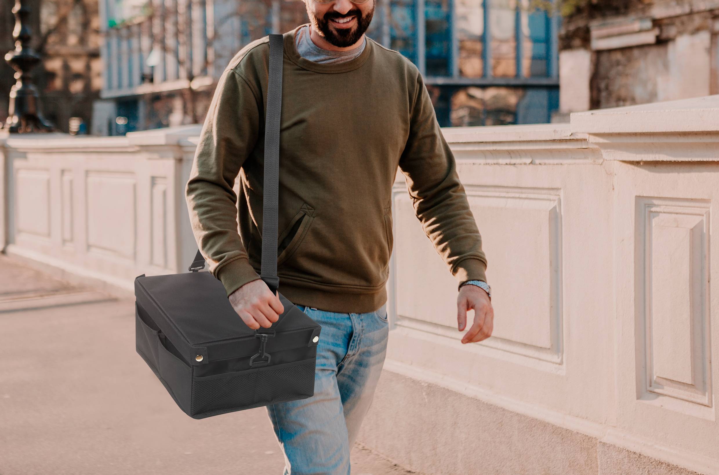 A man is walking along a pavement with a smile, carrying a black shoulder bag. Buildings and trees are visible in the background.