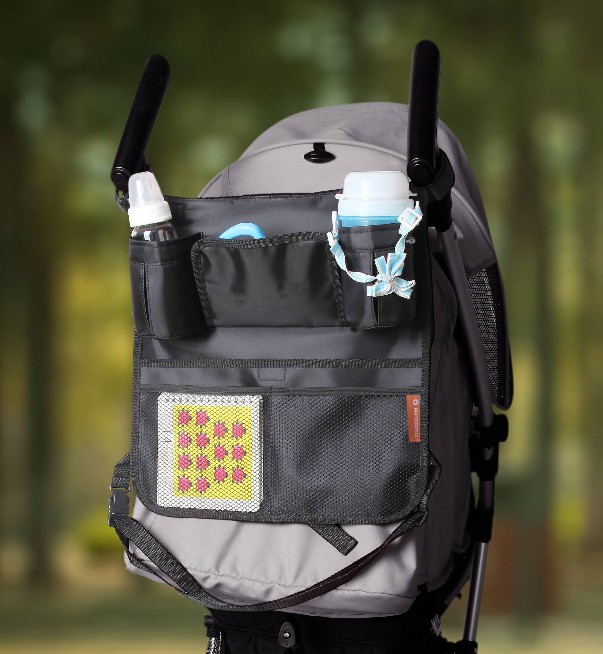 A grey pram bag with bottle holders and a patterned insert is hanging on a pushchair. Background blurred, presumably a park.