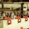 Three orange clamps are holding a wooden board in place in a workshop. In the background, additional blurry clamps can be seen.
