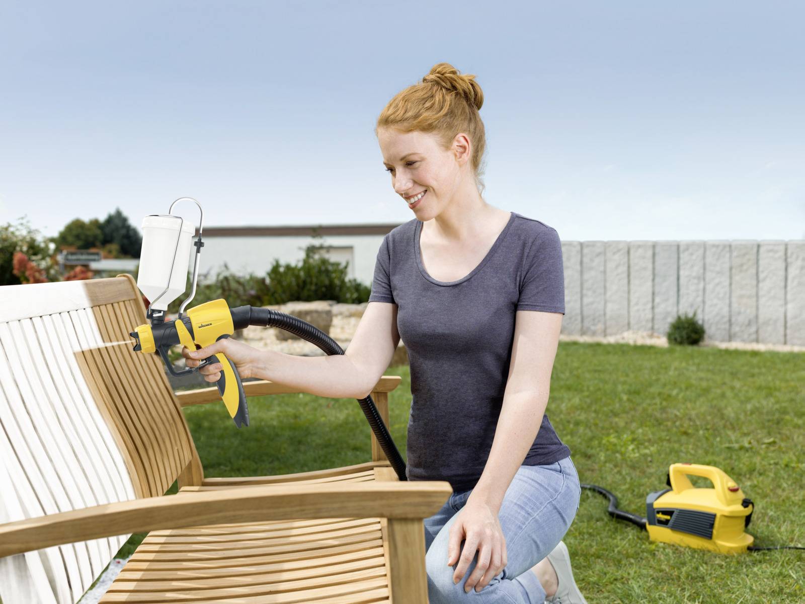 A woman is painting a wooden bench in the garden using a spray painting system. Another device is standing in the grass.