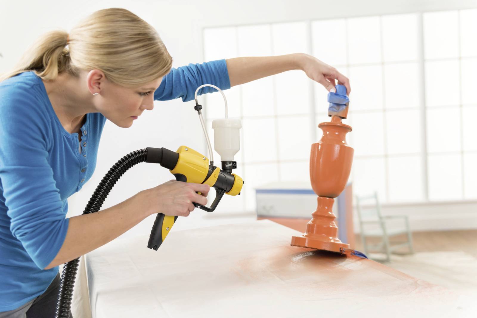 A woman is spray-painting an orange vase with a spray gun. She is focusing on precise work in a well-lit room.