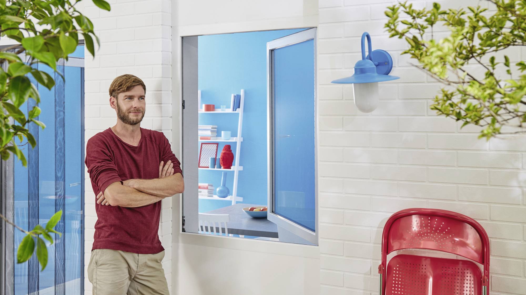 A man wearing a red jumper stands in front of a white building with blue details, a red chair in the foreground, and trees framing the scene.