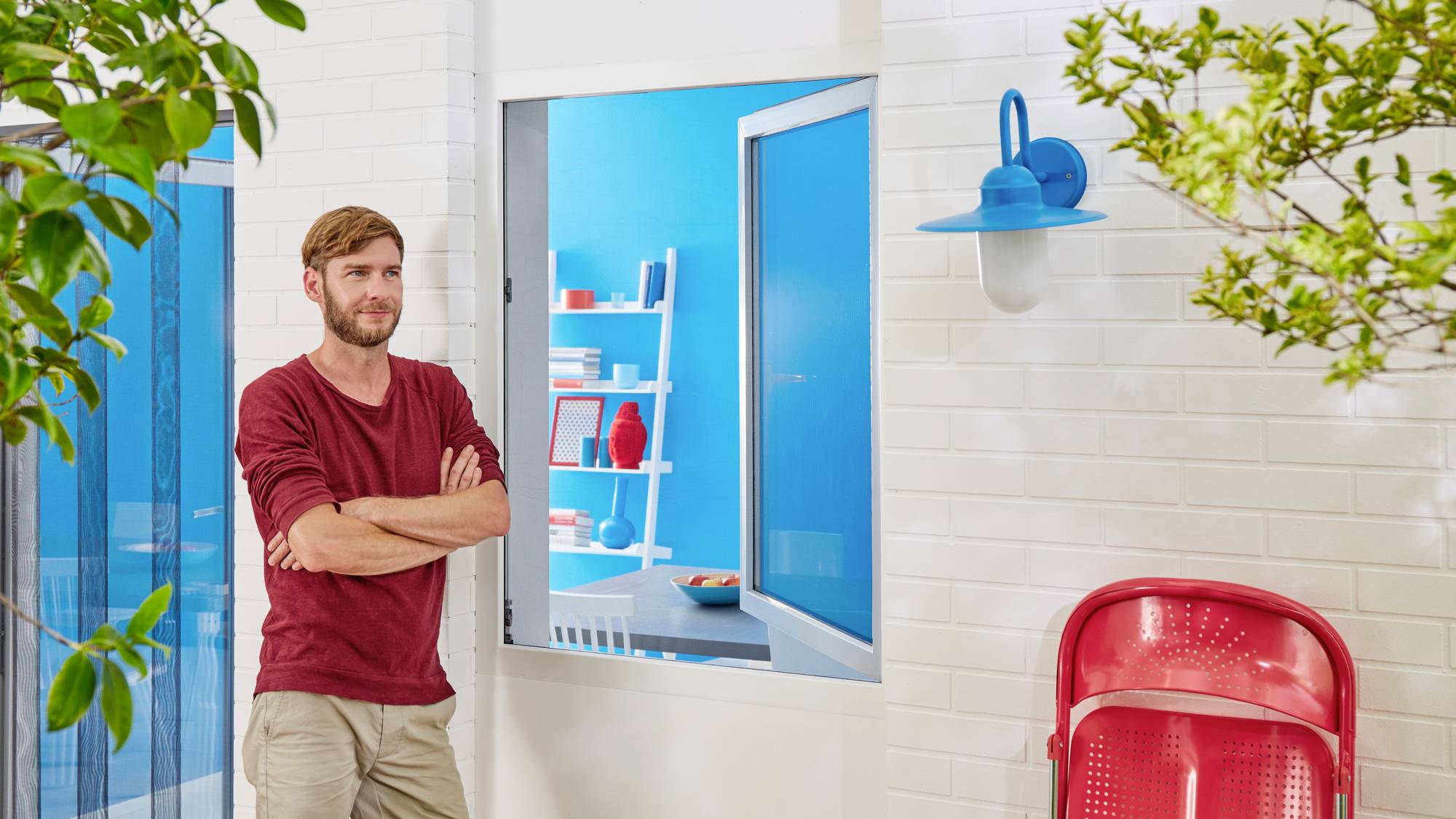 A man is standing outside a brightly painted building with red chairs and an open window, through which a room with blue walls is visible.