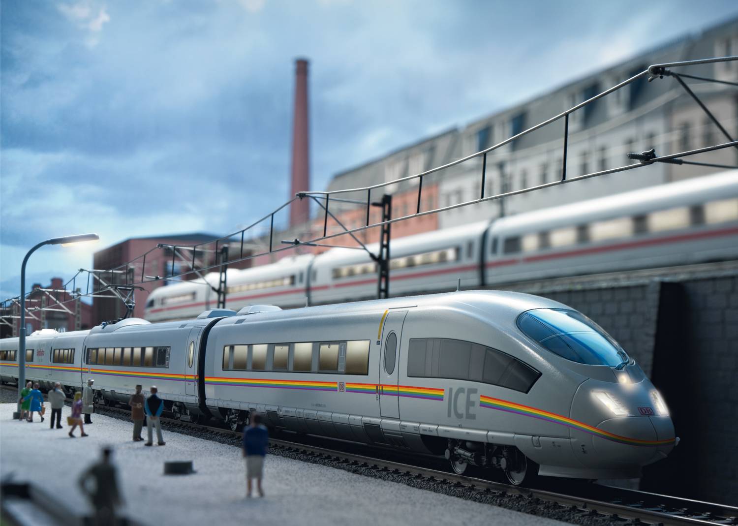 A modern high-speed train on tracks in an urban setting, with people standing on the platform, railway buildings and another train in the background.