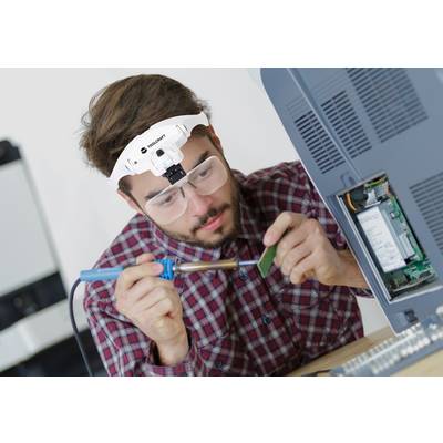A man wearing safety goggles and a head torch is soldering on a computer circuit board with a soldering iron. An open computer sits beside him.