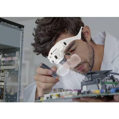 A technician in a white lab coat is working intently on an open computer circuit board. He is wearing a magnifying lens to aid his precision.