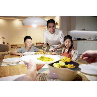 A man is serving dinner to a family at a table in a cosy kitchen. Two children are waiting with smiles for the food.