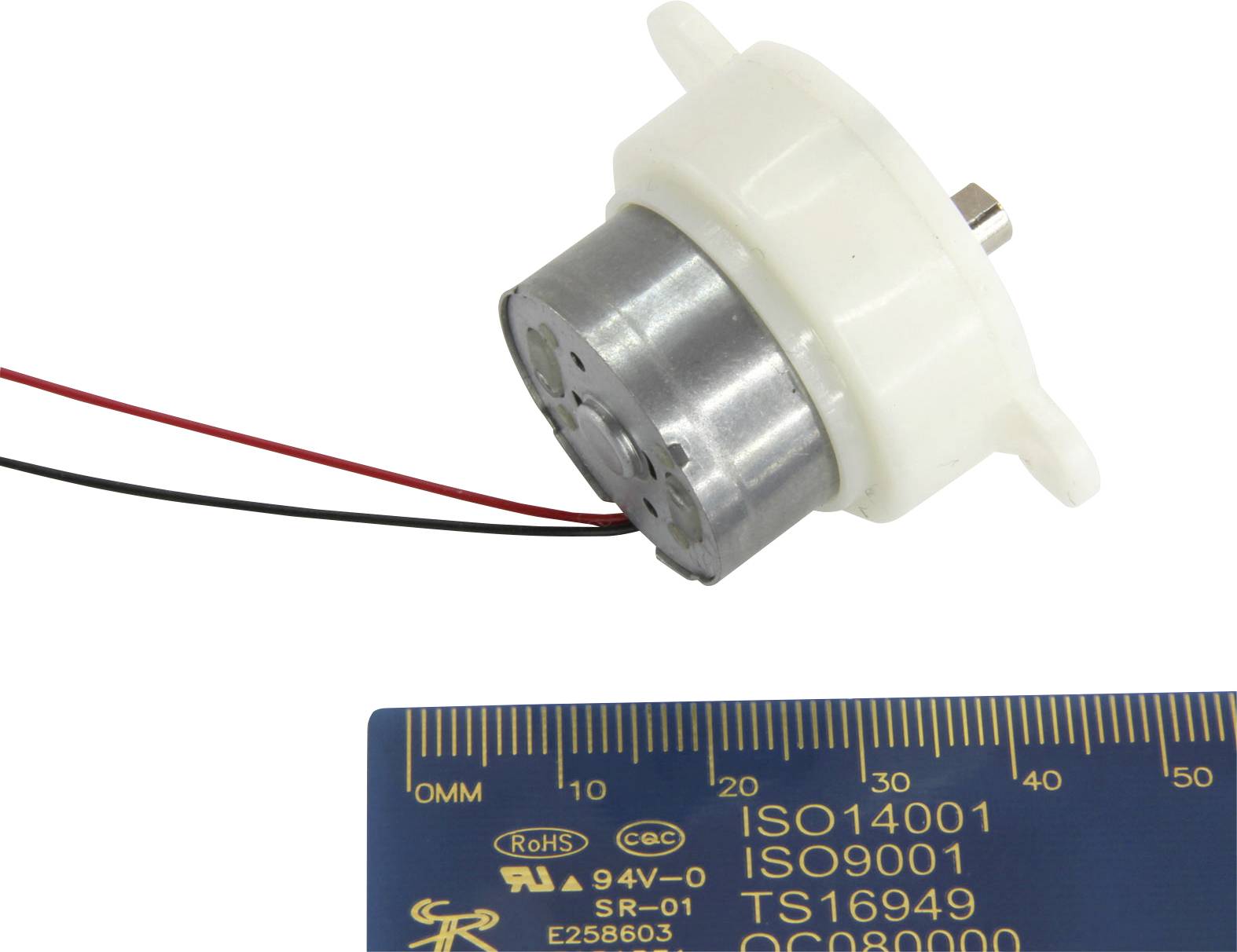 A small electric motor with a plastic casing and two protruding wires lies next to a ruler showing metric measurements.