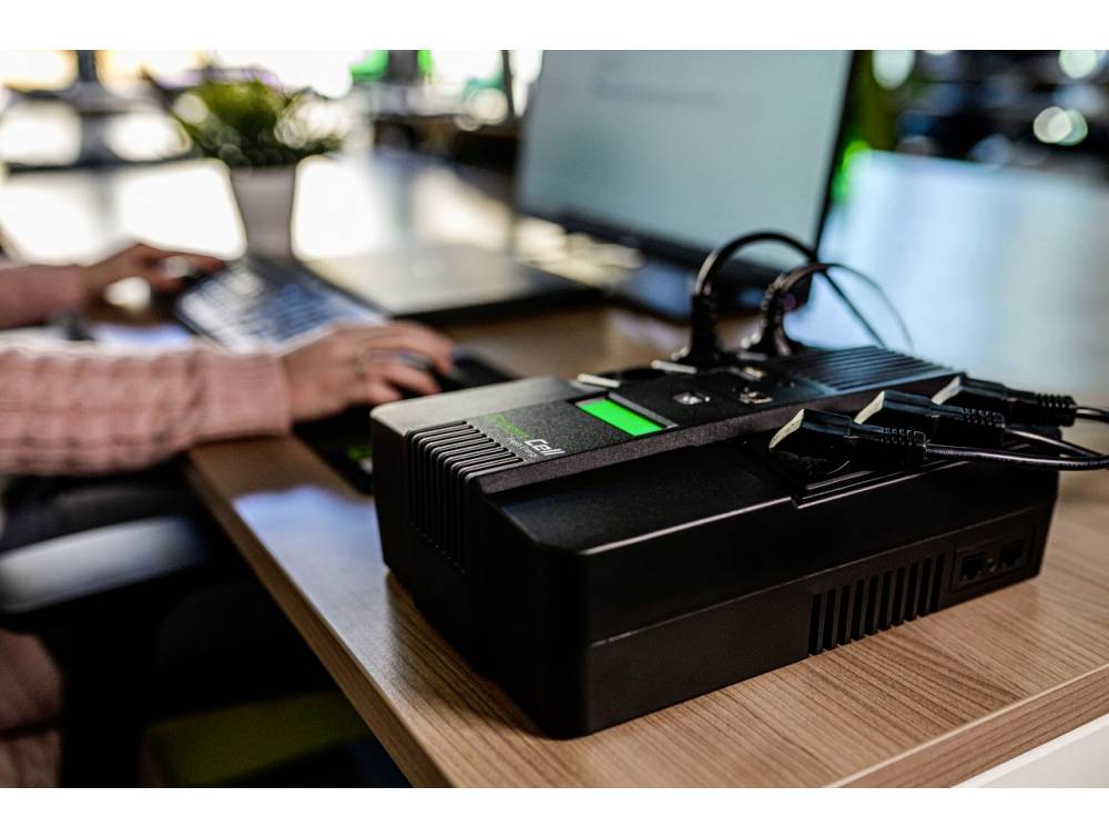 A person is working on a laptop. In the foreground, there is an electronic device with cables. This represents a technical office setup.