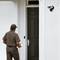 A delivery driver stands in front of a front door holding a parcel. A security camera is mounted above the door.