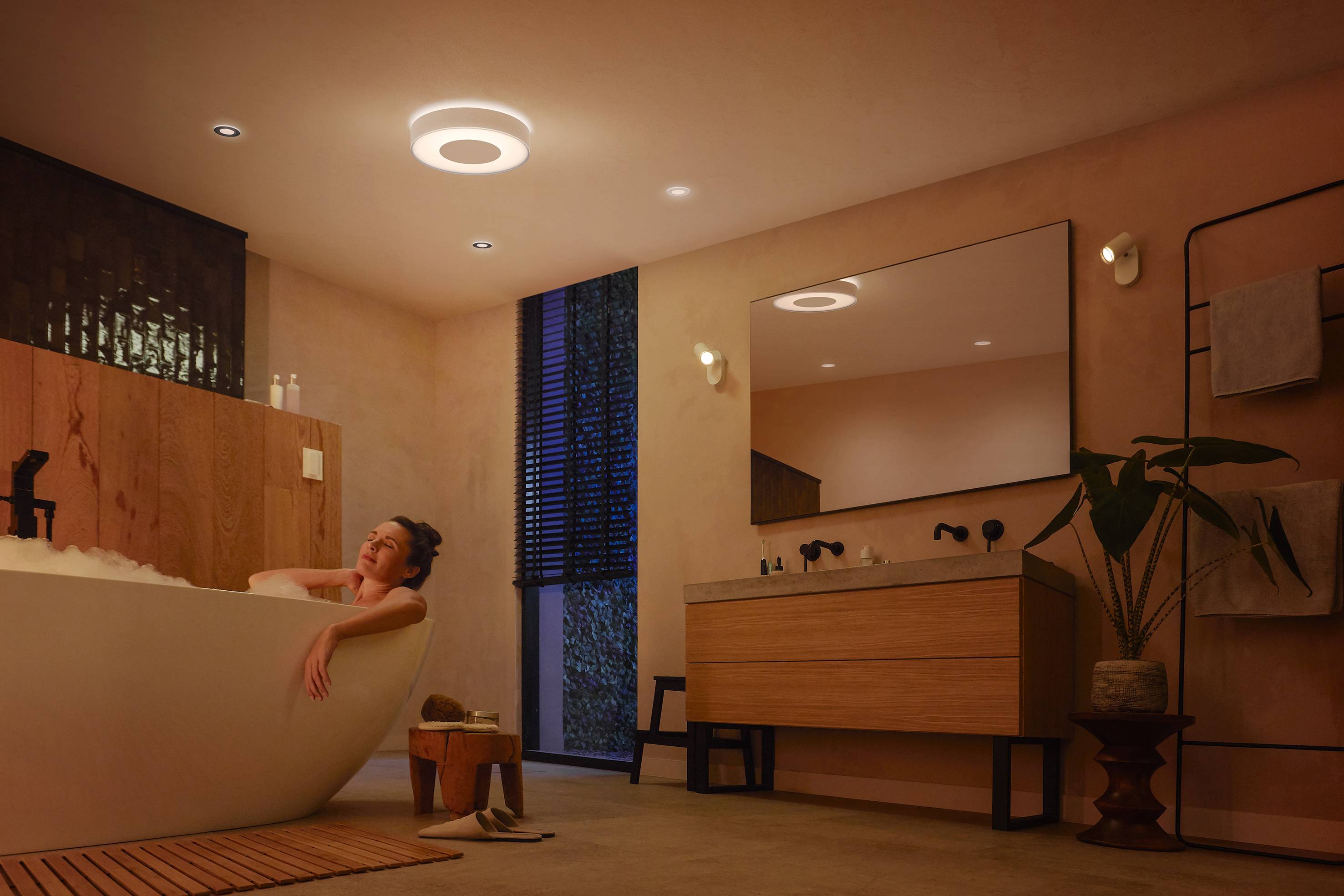 A person is relaxing in a modern bathroom with dimmed lighting, lying in a white bathtub, decorated with plants.