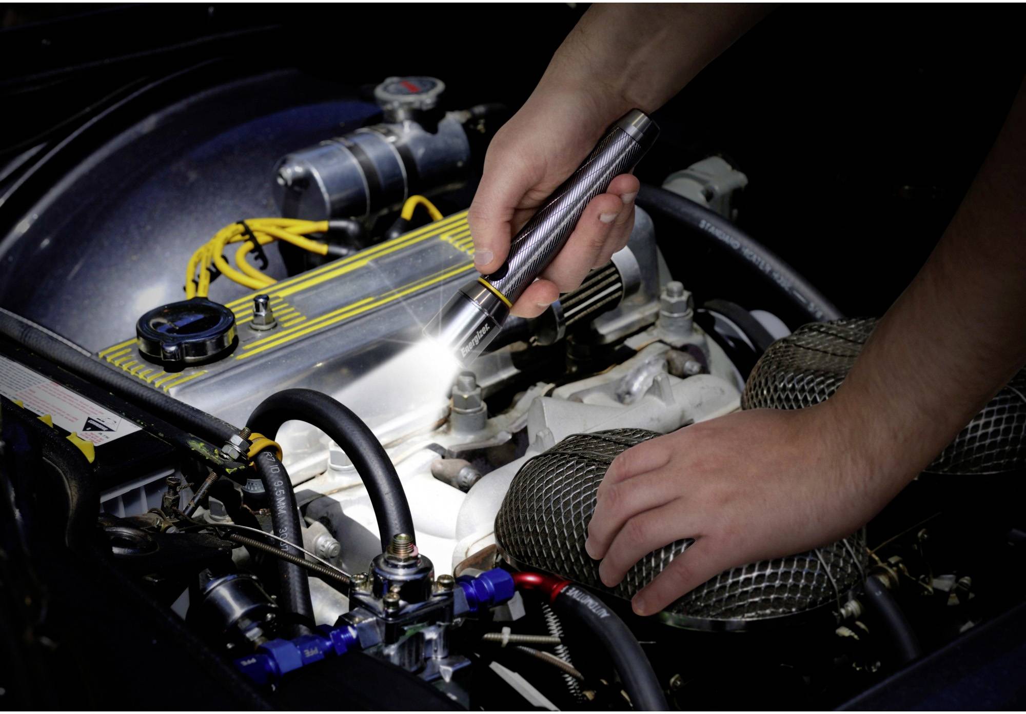 A person is examining an open car engine with a torch, surrounded by cables and hoses.