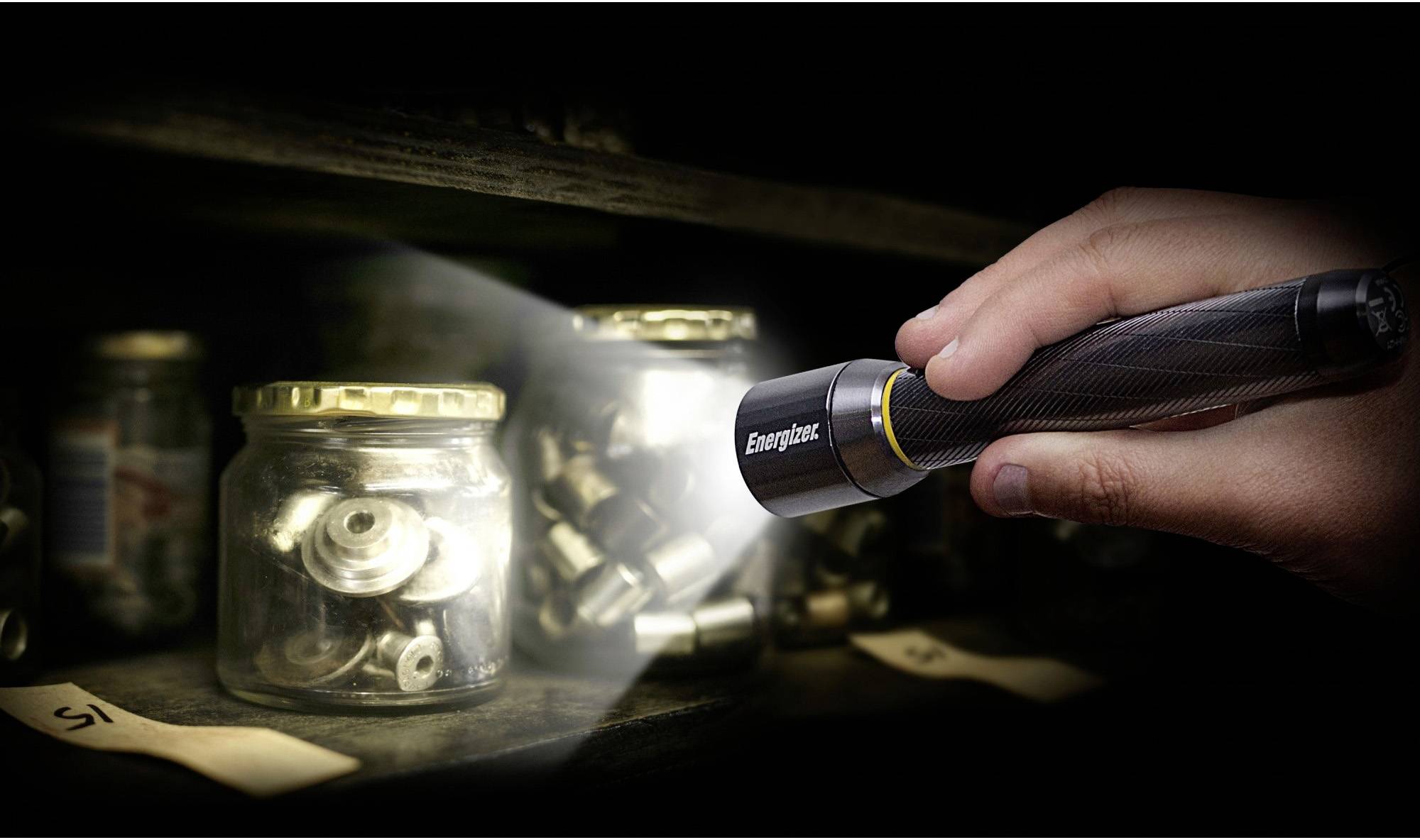 A hand holds a torch and shines light on glass containers with metal parts in a dark room or warehouse.