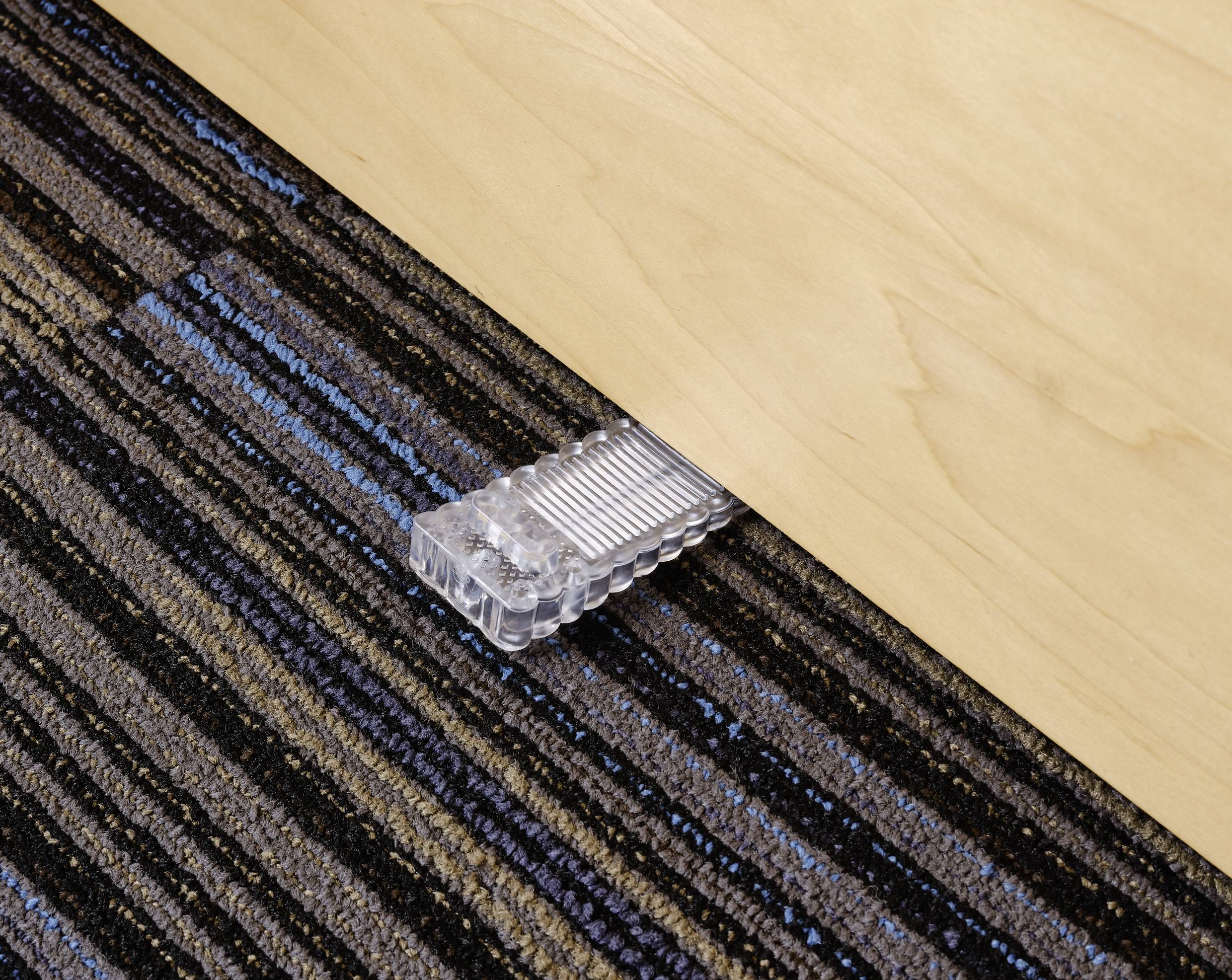 A door wedge holds a wooden door open. The door wedge is transparent and is resting on a striped carpet.