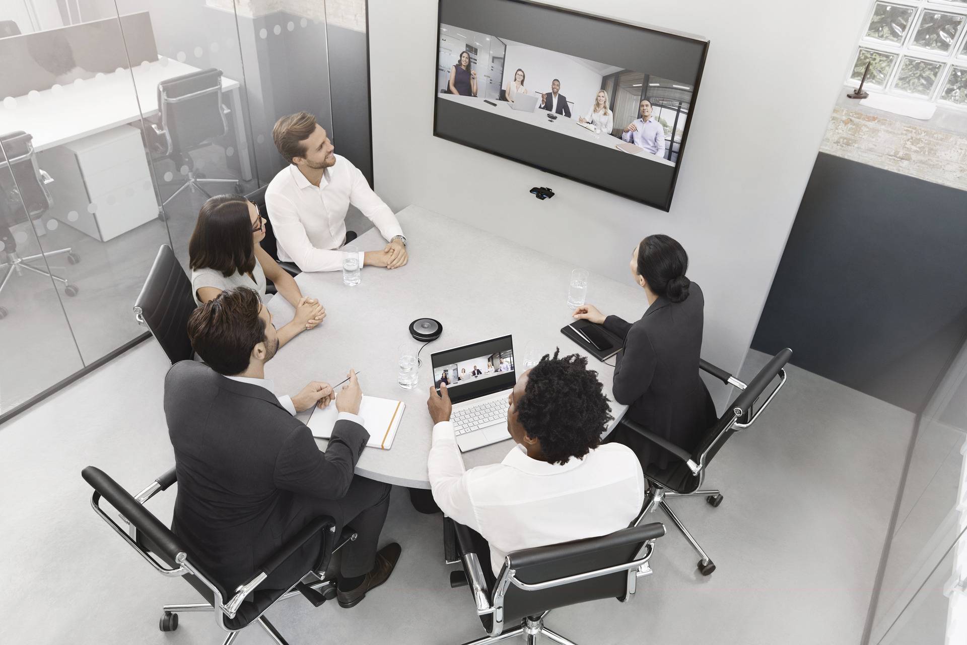 Five people are sitting in a conference room conducting a virtual meeting with participants on a large screen.