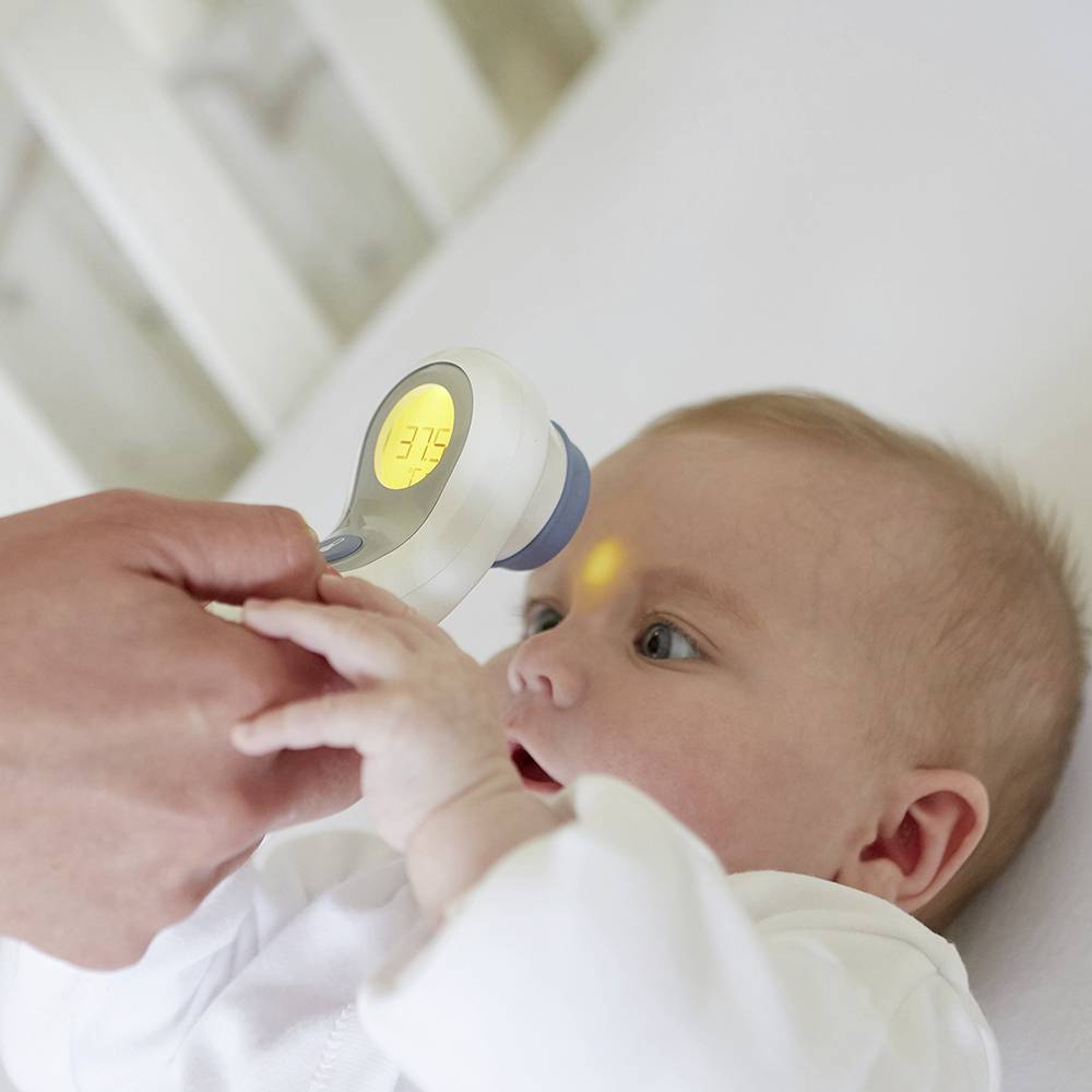 An adult is measuring a baby's temperature using a forehead thermometer. The display shows 37.5 degrees Celsius.
