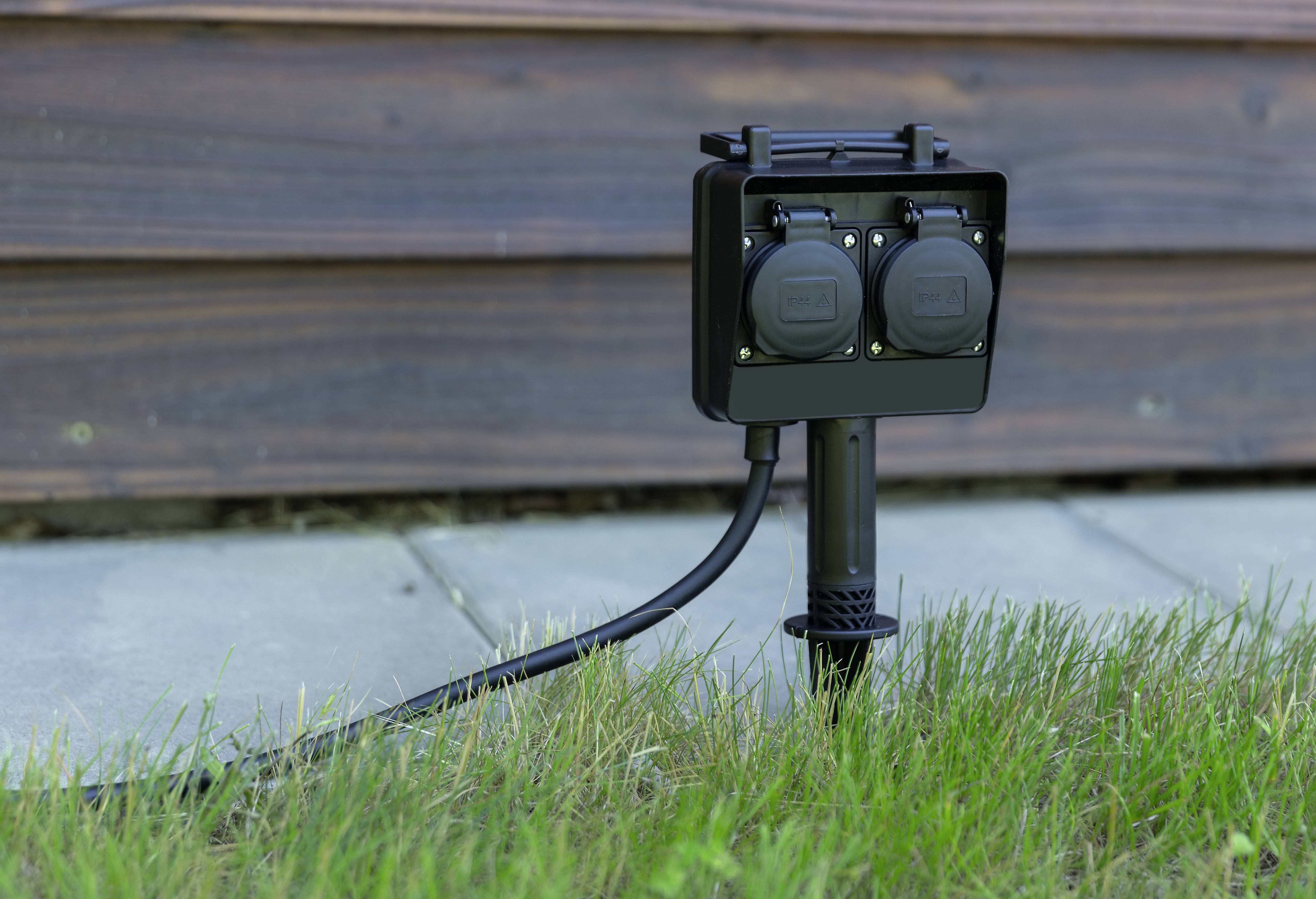 A black outdoor socket with two connections is mounted on a metal post, situated in the grass near a wooden building.
