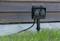 A black outdoor socket with two connections is mounted on a metal post, situated in the grass near a wooden building.