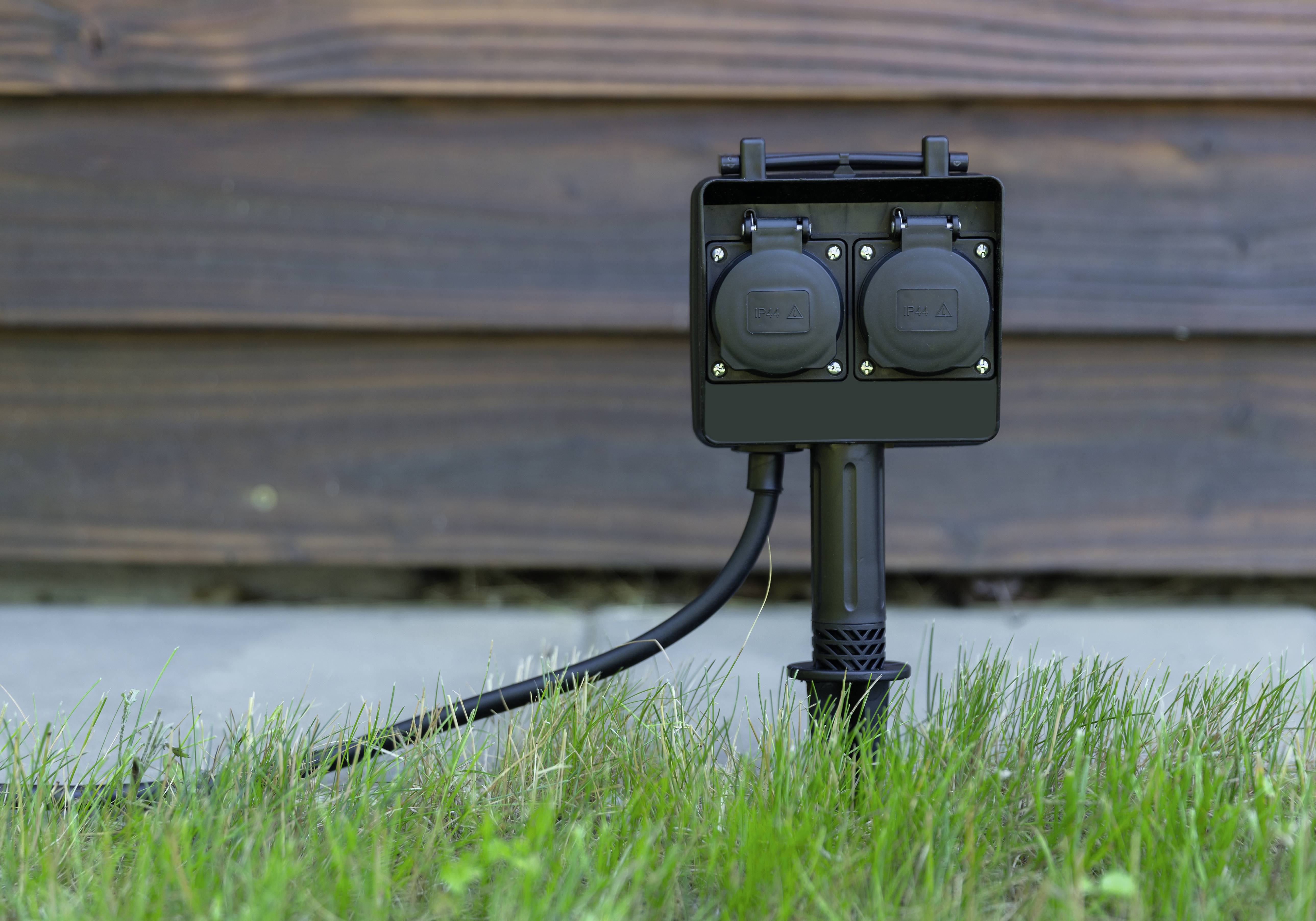 A black outdoor socket with two outlets is installed on a lawn. A wooden wall can be seen in the background.