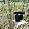 A black rain gauge stands on a white table outdoors, surrounded by green plants.