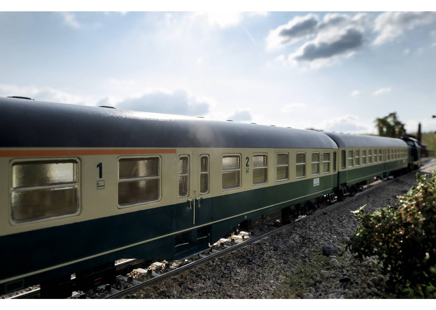 A model train travels along a track through a landscape with a blue sky and a few clouds. The carriages are painted beige and blue.