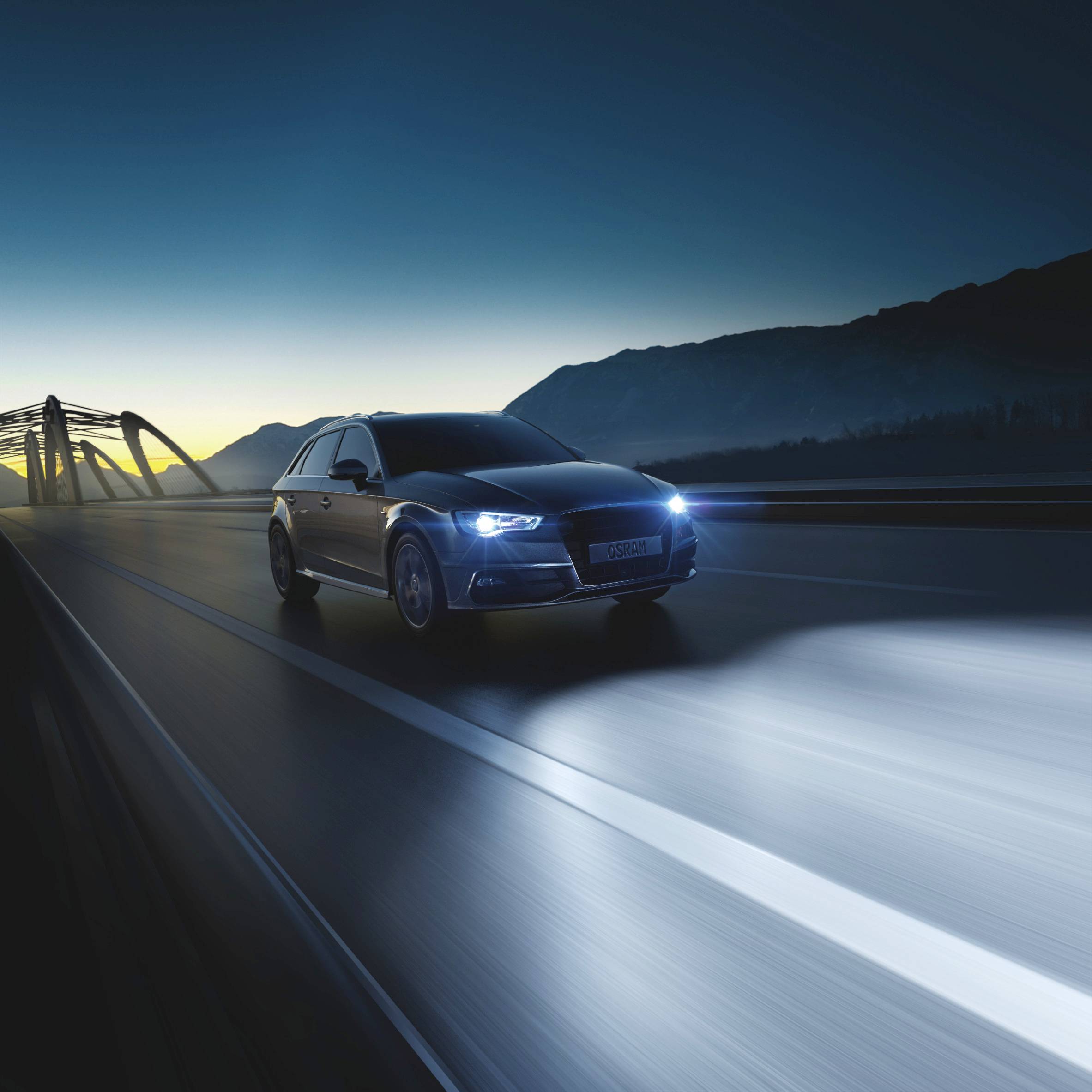 A car drives at night across an illuminated bridge, surrounded by hills. The sky is dark blue, and the bridge has a futuristic design.