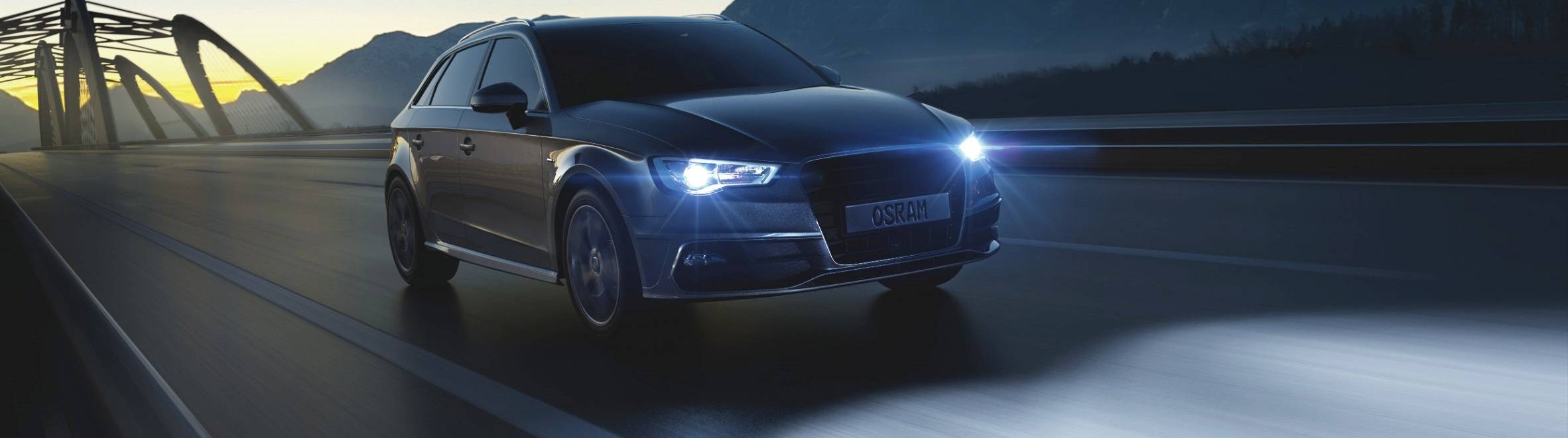 A car drives across a bridge at night, its headlights illuminating the way. Mountains and a softly lit sky are visible in the background.