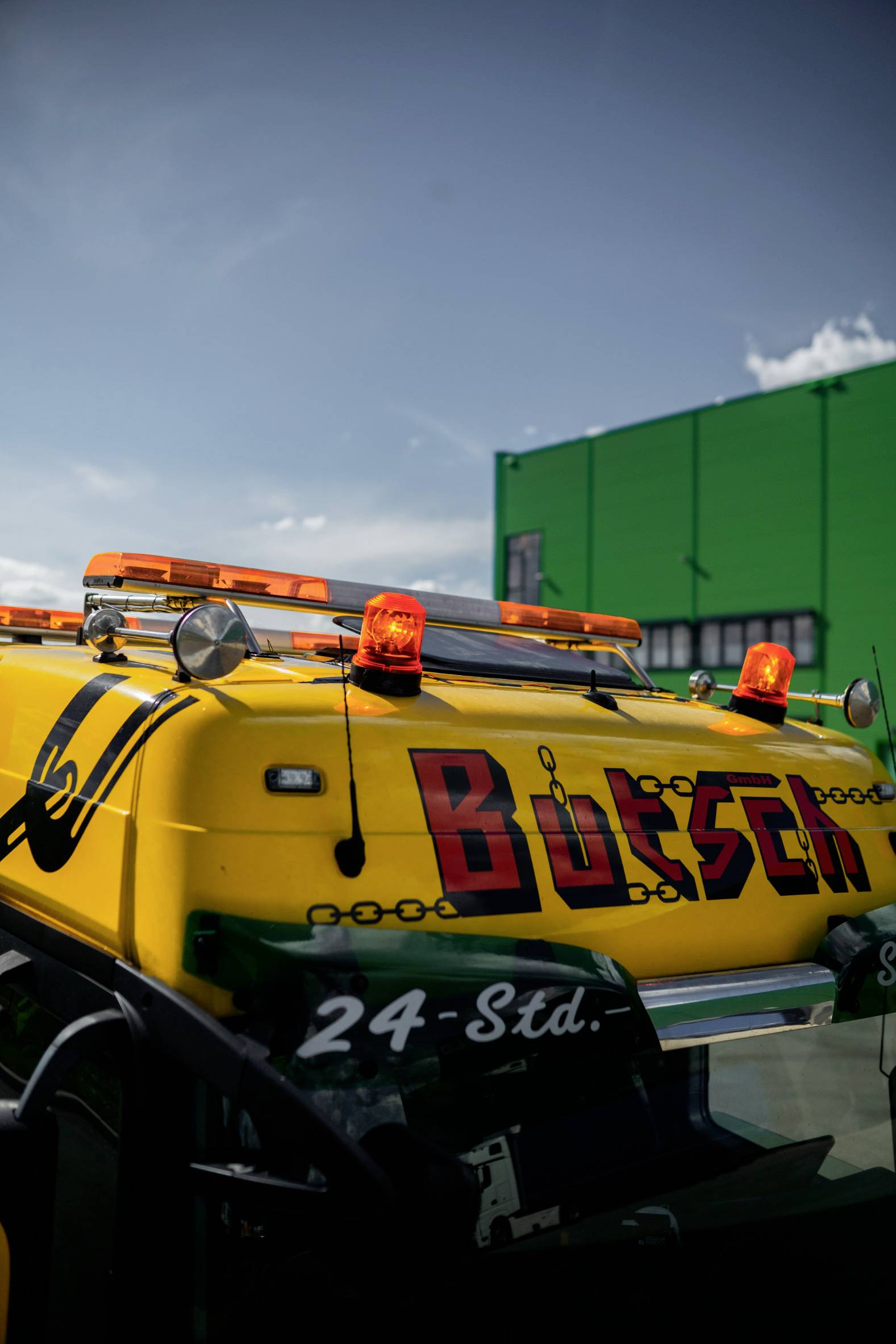 A yellow recovery vehicle with orange warning lights and the 'Bösch' lettering on its side, positioned in front of a green building.