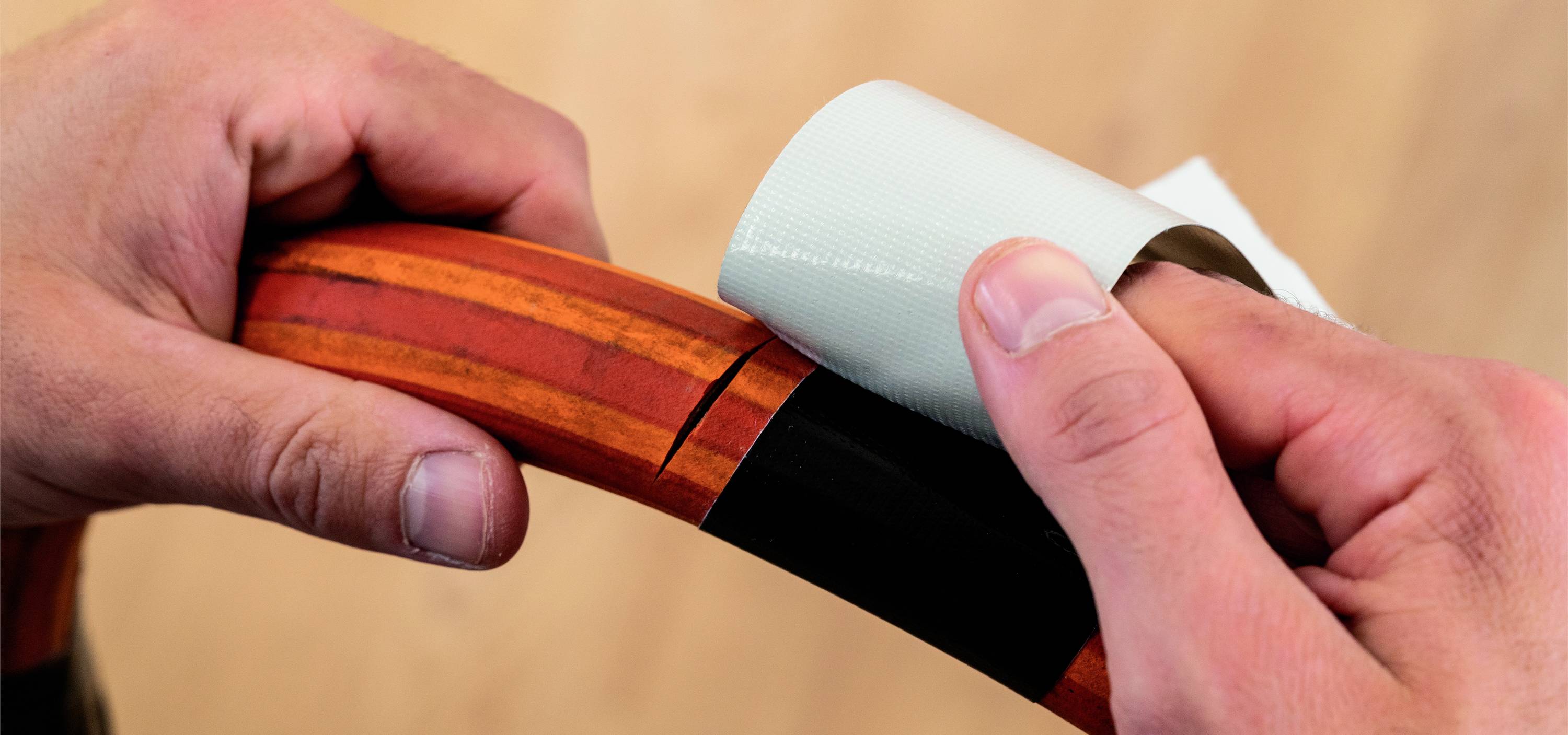 Two hands are wrapping a piece of white tape around a wooden handle, likely for repair or reinforcement of a tool.