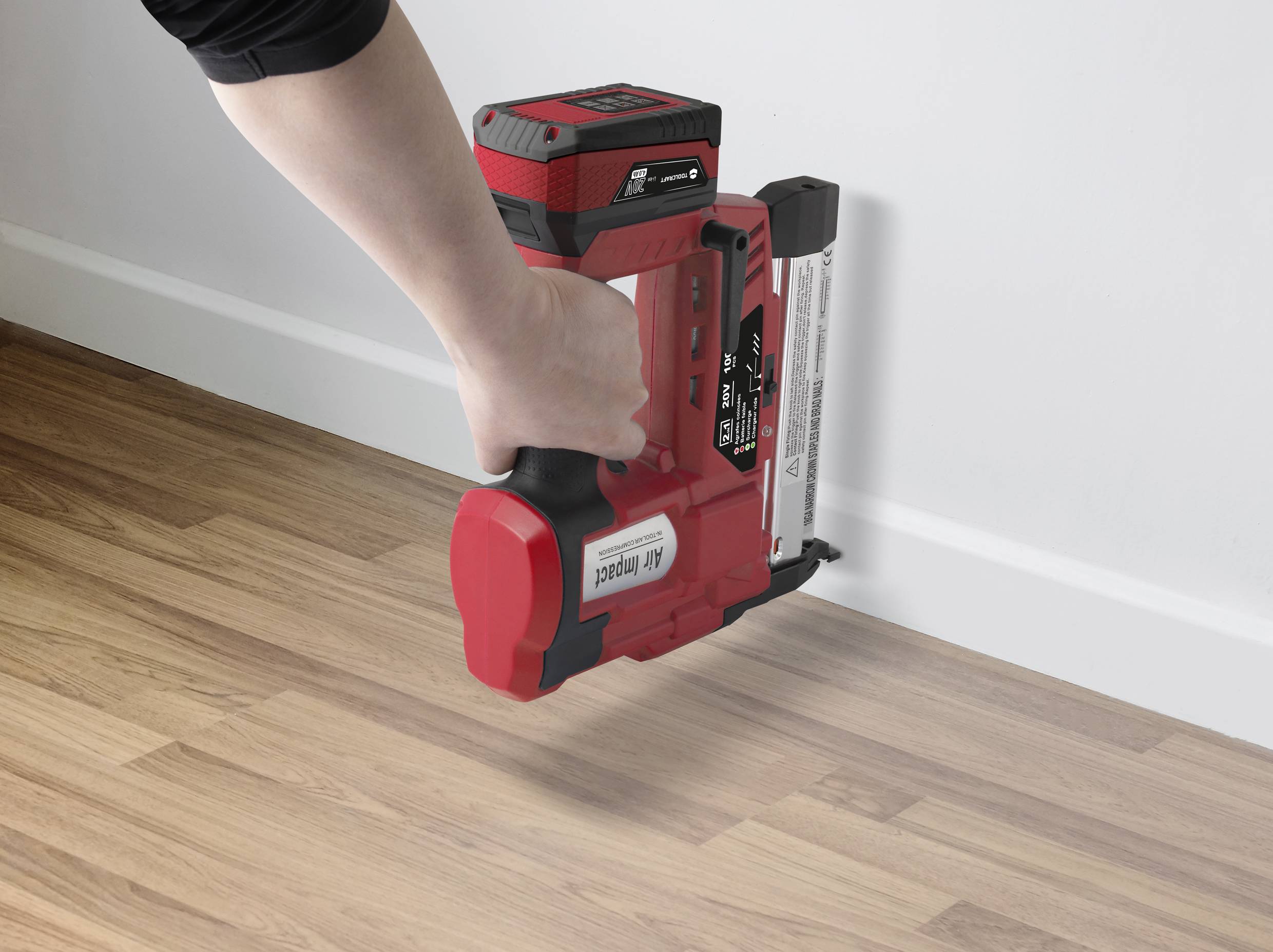 A hand is holding an electric nail gun, firing a nail into the wall above a wooden floor. The device is red and black.
