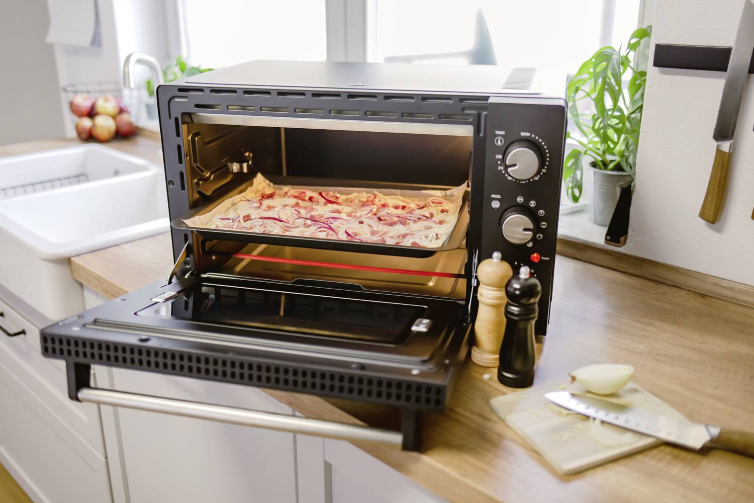 A pizza oven on a kitchen worktop, with a rectangular pizza currently baking inside. Salt and pepper mills are positioned nearby.