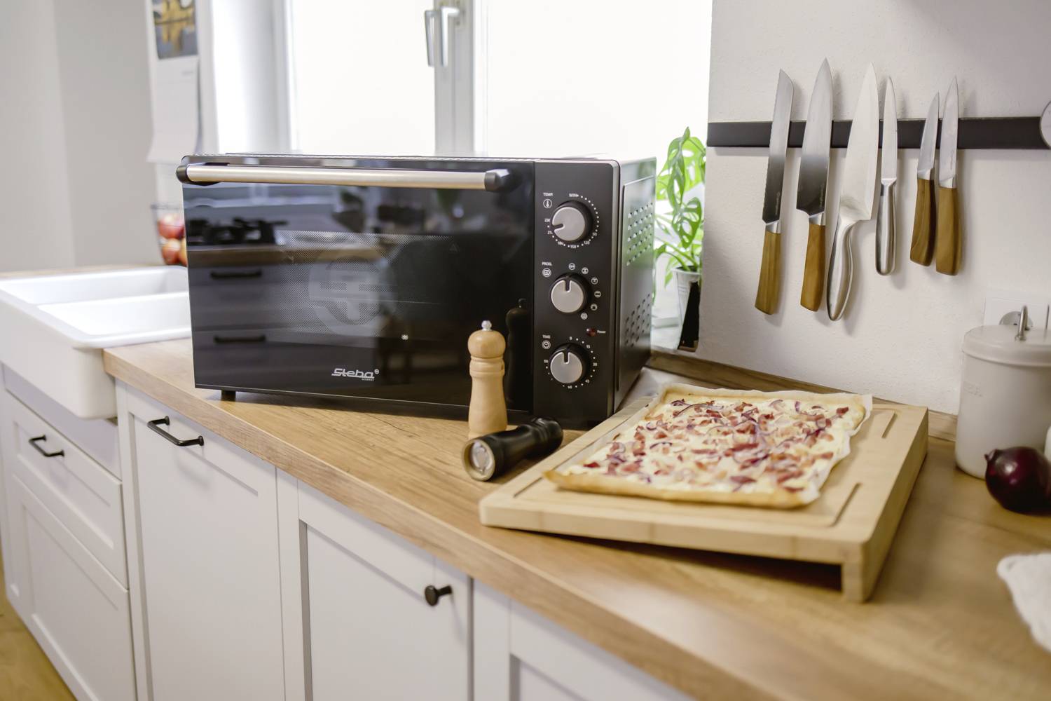 A kitchen with a small, black oven on a wooden worktop. Beside it lies a tarte flambée on a wooden board.