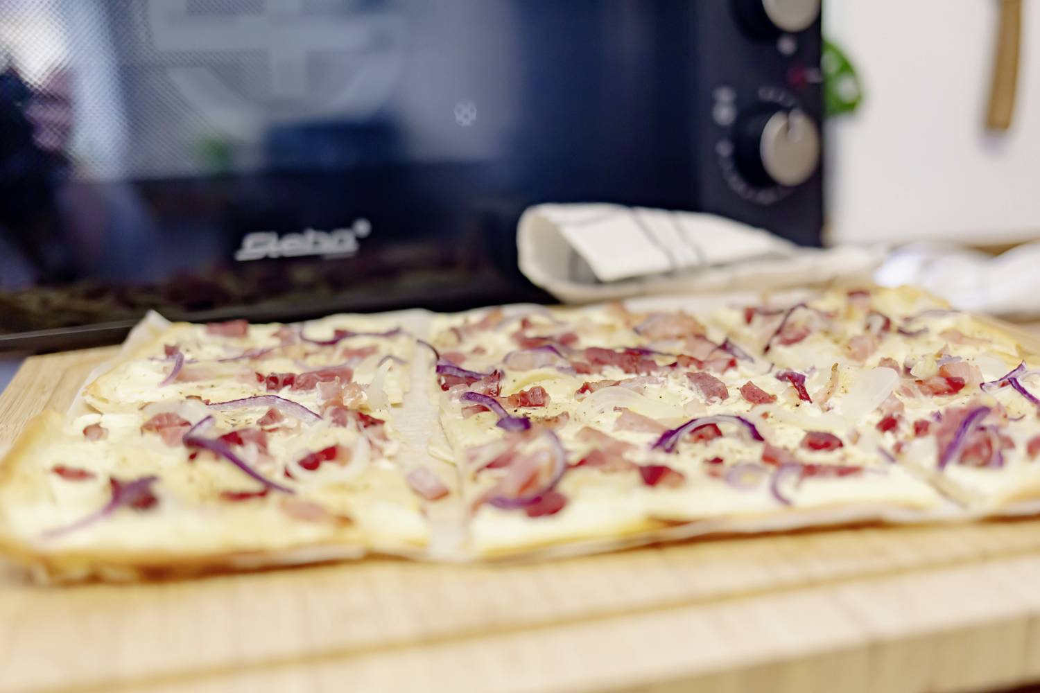 A freshly baked tarte flambée lies on a wooden board in front of an oven. Topped with onions and bacon.