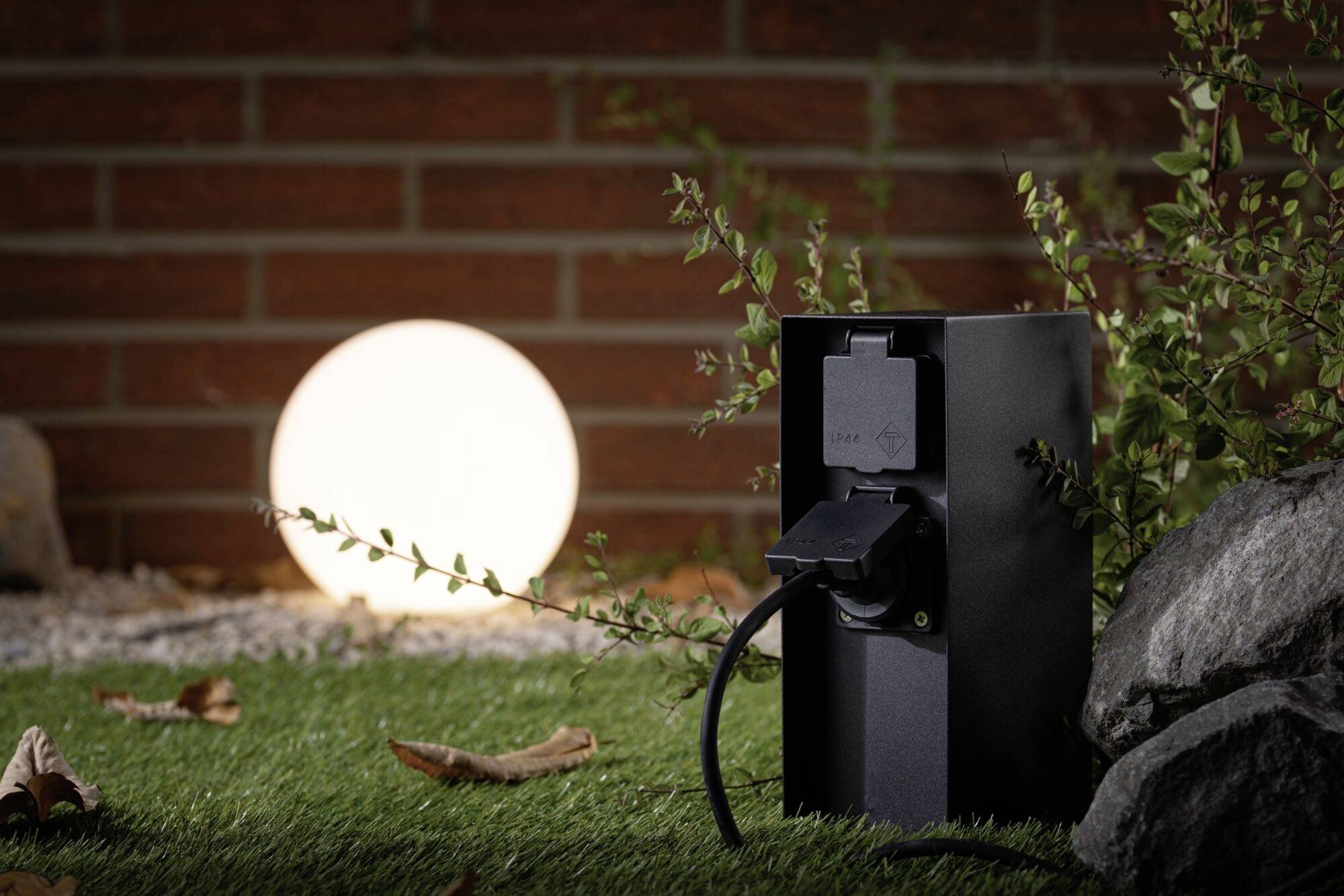 An outdoor socket stands on a lawn area in darkness; a round lamp glows in the background, surrounded by leaves.