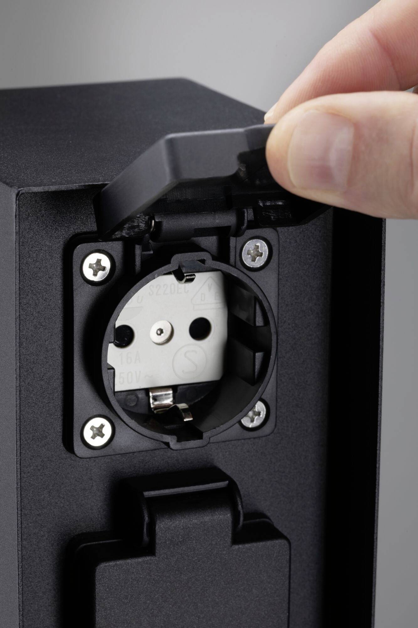 An open socket on a black casing. A hand lifts the socket cover.