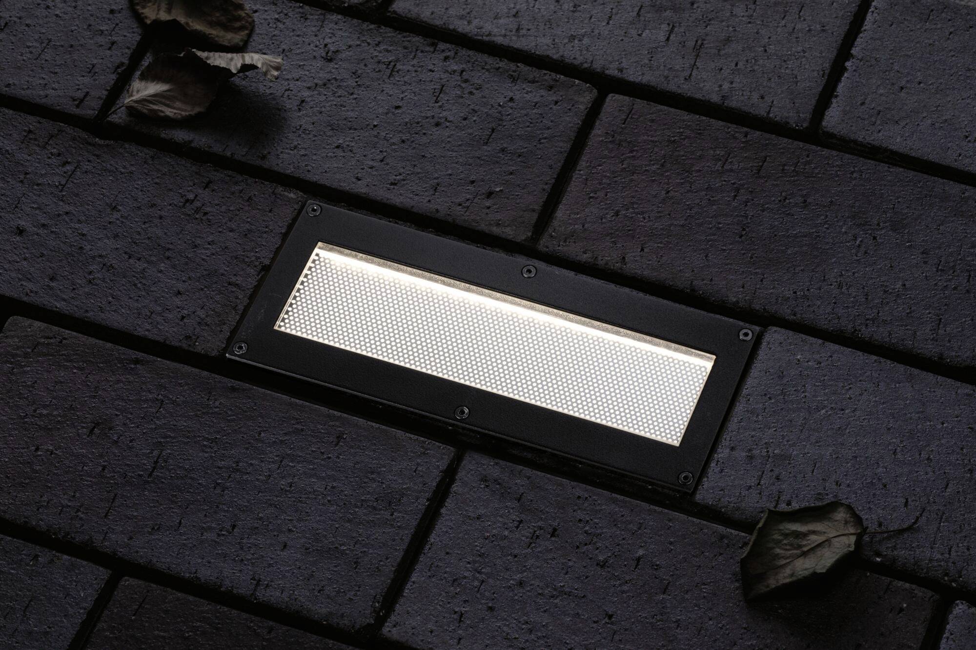 A rectangular floor light embedded in dark paving stones dimly illuminates the surroundings. Two dry leaves lie beside it.