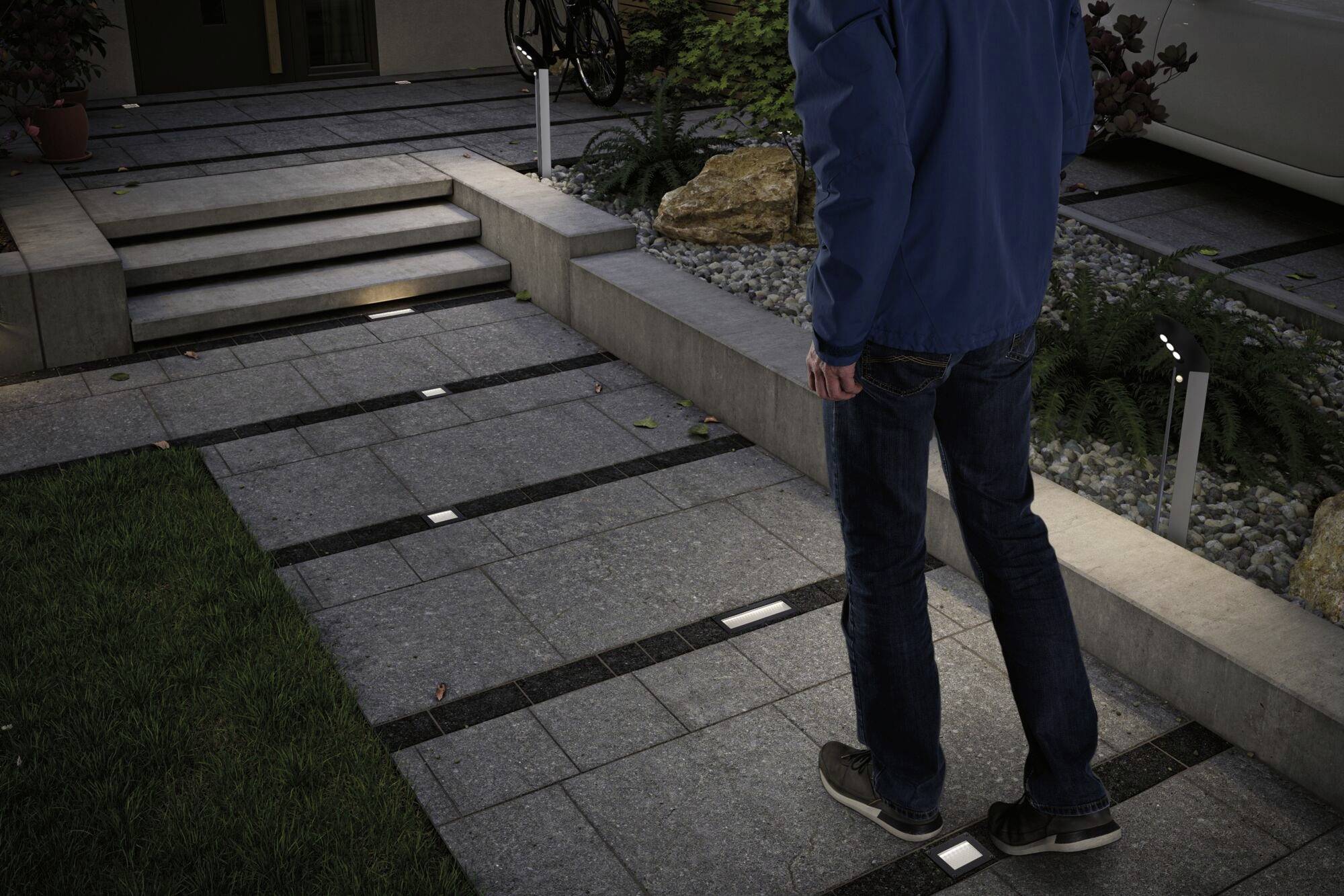 A person is walking on an illuminated pathway made of grey tiles. Steps lead to an elevated entrance. Plants and rocks line the path.