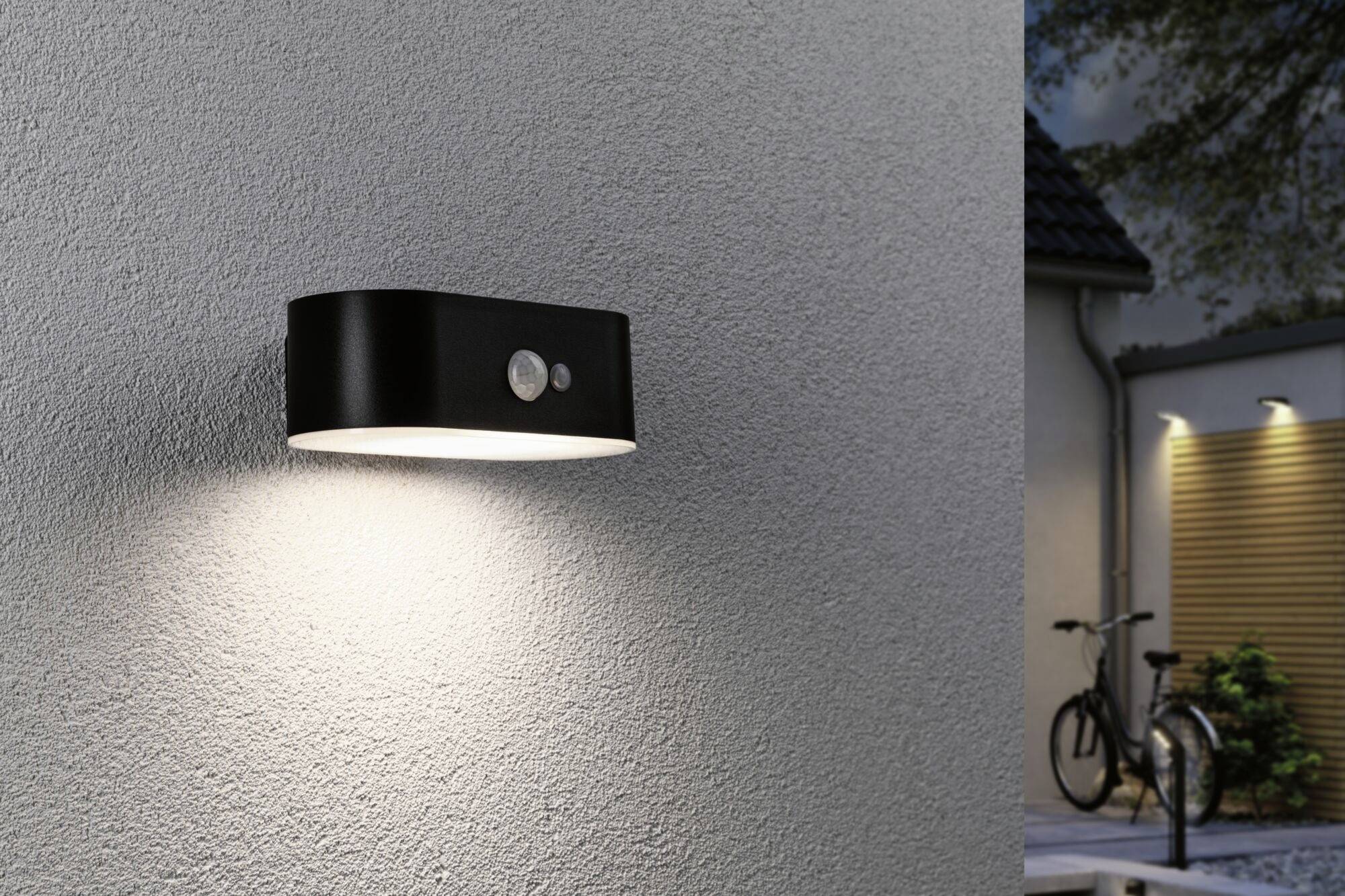 A black outdoor wall light with a motion sensor illuminates a grey wall. In the background, a lit courtyard can be seen.