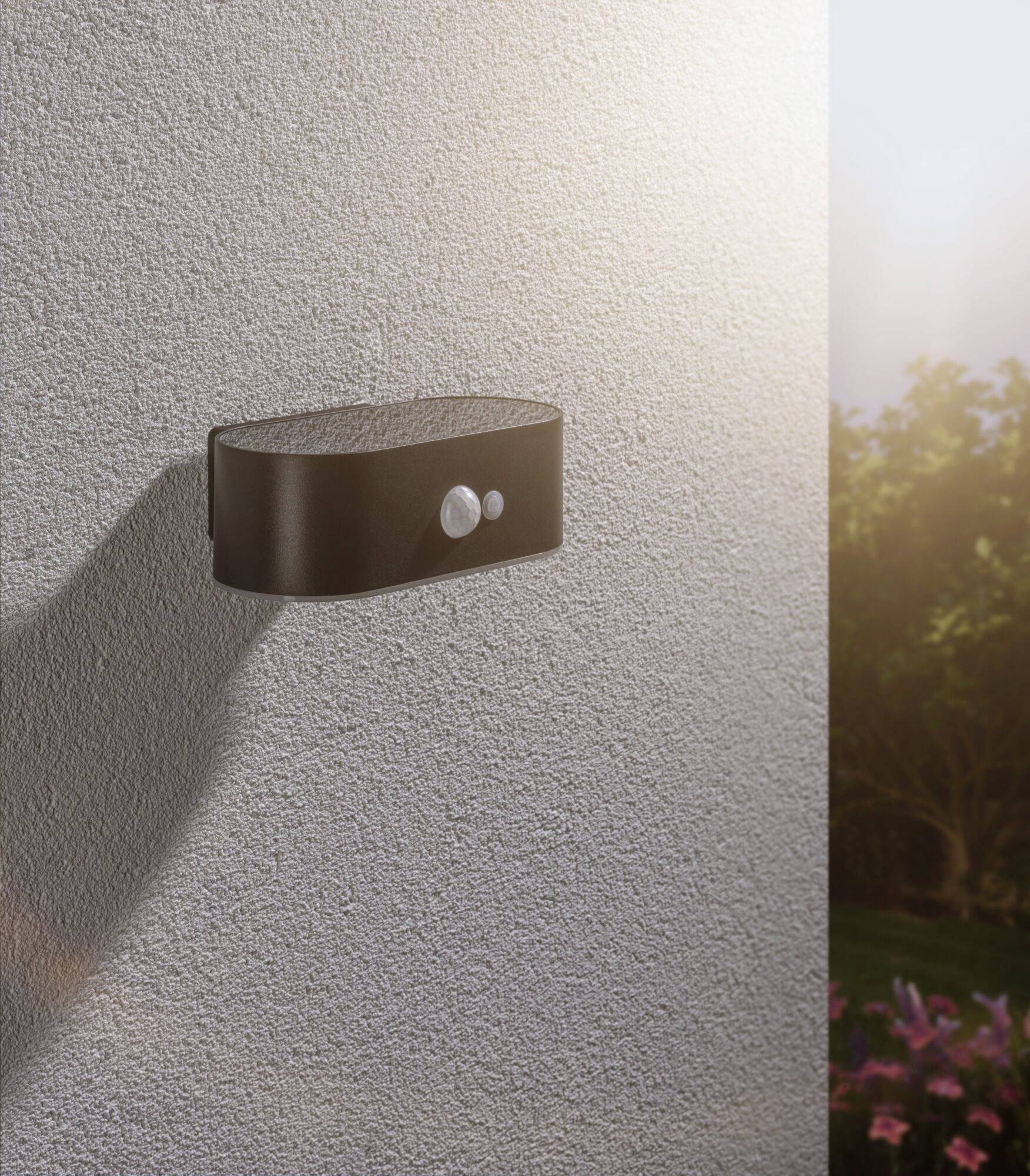 Outdoor light with motion sensor on a house wall, sunny sky in the background, illuminating garden area in darkness.