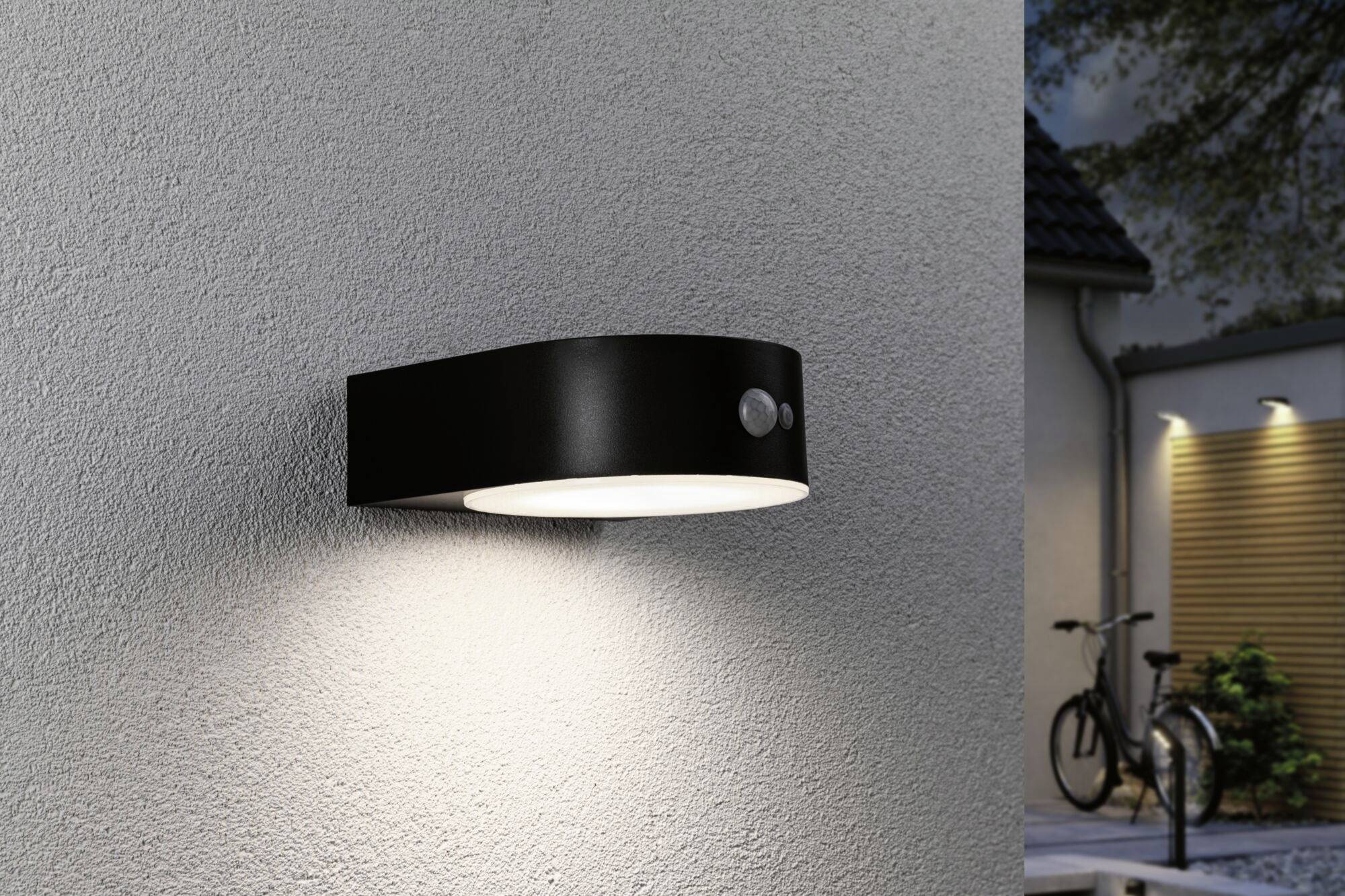 Black outdoor lamp on a house wall, shining brightly. A blurred house and bicycle in the background.