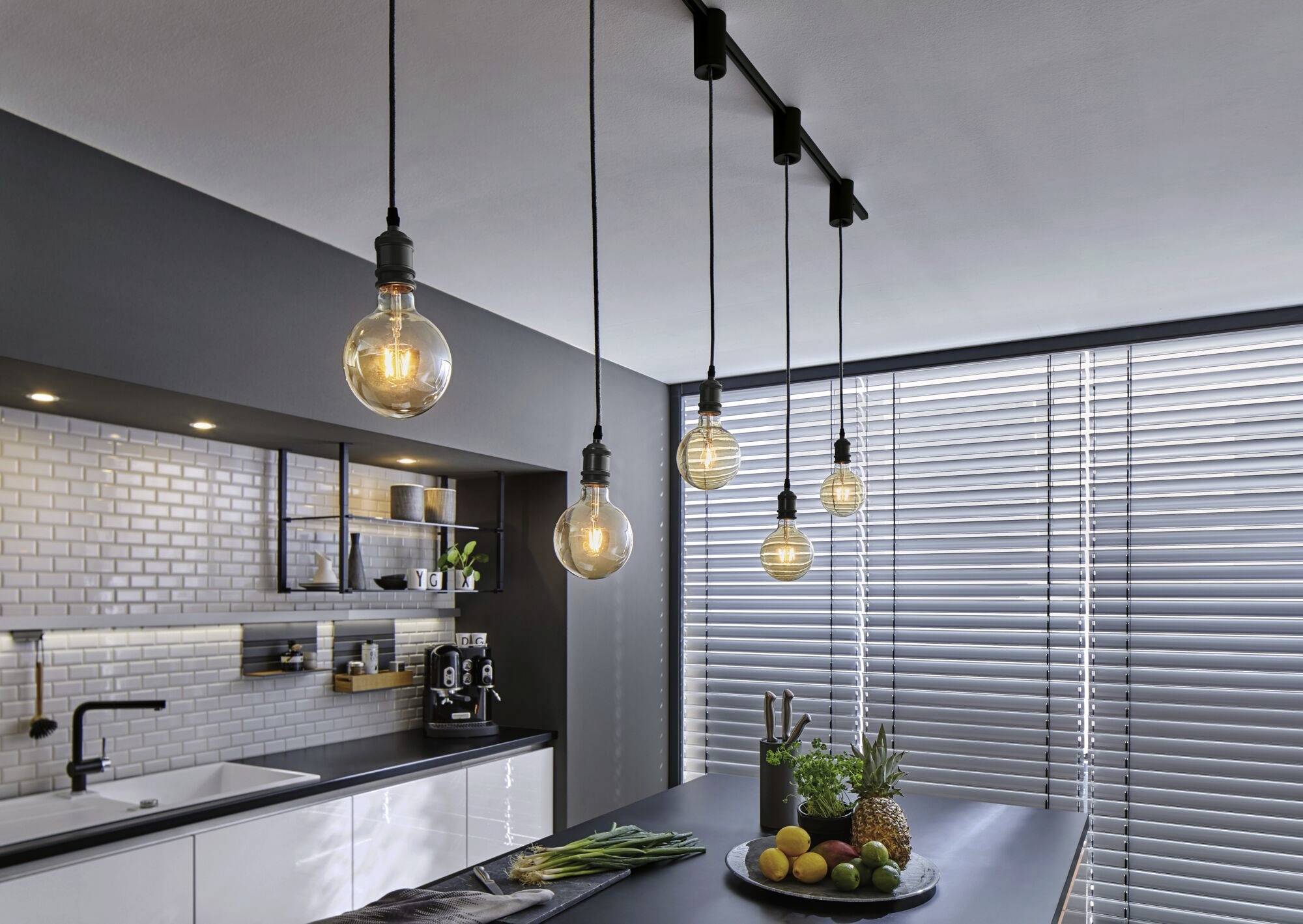 A modern kitchen with hanging light bulbs, a fruit bowl on the island counter and large venetian blinds on the window.